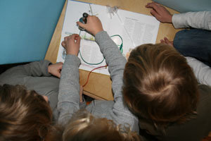 vor dem Anleitungsbuch wird ein mit Kabel verbundenes Dynamo von Kindern betrachtet